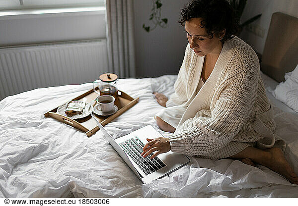 a woman in a sweater is sitting on the bed and having breakfast