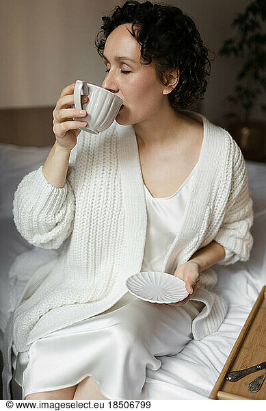 a woman in a sweater is sitting on the bed and having breakfast