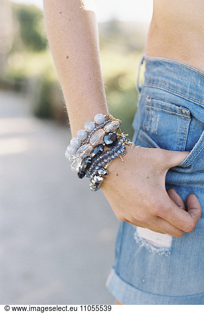 A woman in denim shorts and a bare midriff. A thumb hooked into her pocket  and wrist with bracelets.