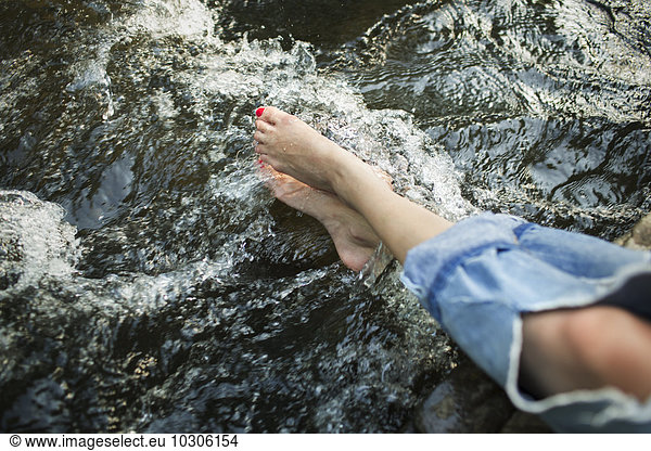 A woman in fashionable jeans with a rip  with her feet in the cool flowing waters of a river.