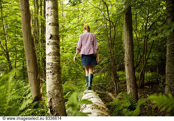 A woman in wellingtons walking along a fallen tree trunk  in woodland.