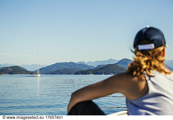 A woman looks over a calm sea scene with sailboat and mountains