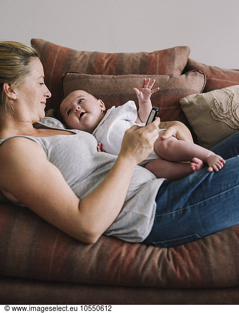 A woman lying on a sofa holding a baby girl  and holding a smart phone in one hand.