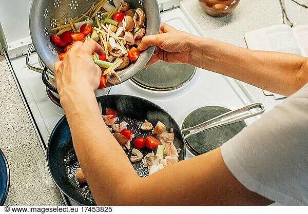 A woman pours chopped vegetables into a frying pan on the stove