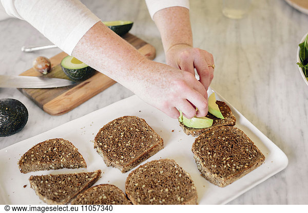 A woman preparing a sandwich with slices of brown bread and sliced avocado.