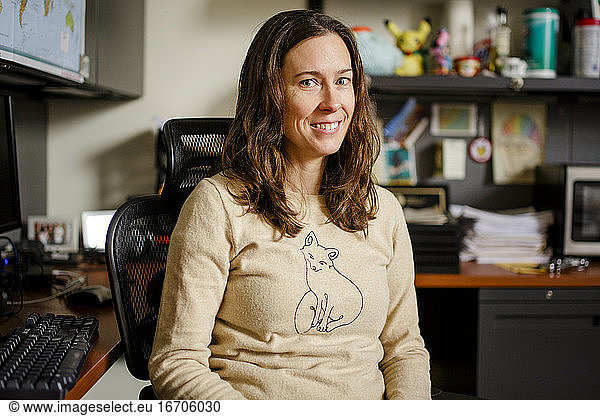 A woman professor sits at her office desk smiling with direct gaze