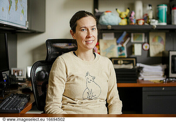 A woman professor sits in her office at a desk smiling