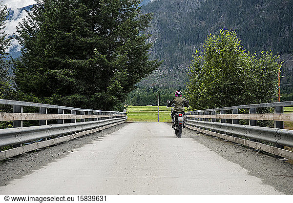 A woman rides her motorcycle across a bridge on a cloudy summer day.