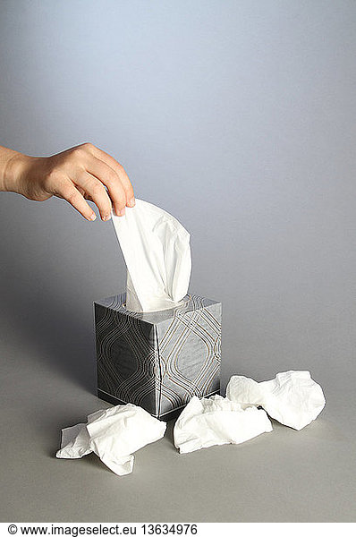 A woman's hand removing a tissue from a tissue box. Used  crumpled tissues surround the tissue box.