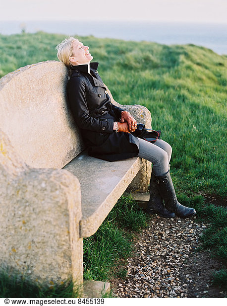 A woman seated on a stone bench on a coastal path.