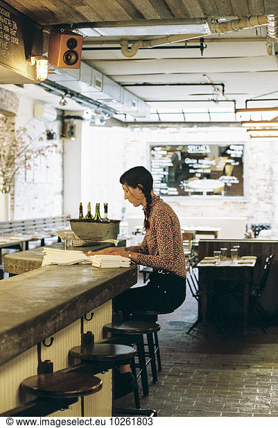 A woman seated on a tall stool  folding napkins for a restaurant table.