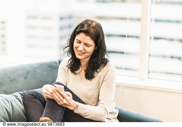 A woman seated with her feet up on a sofa  looking at her smart phone.