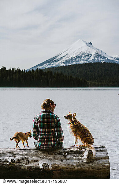 A woman sits and enjoys view of lake and mountains with her two dogs