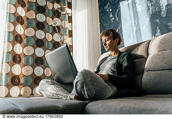A woman sits comfortably on the couch  legs crossed  and uses a laptop