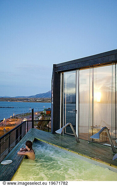 A woman sits in an empty hot tub overlooking the waters surrounding a small coastal city.