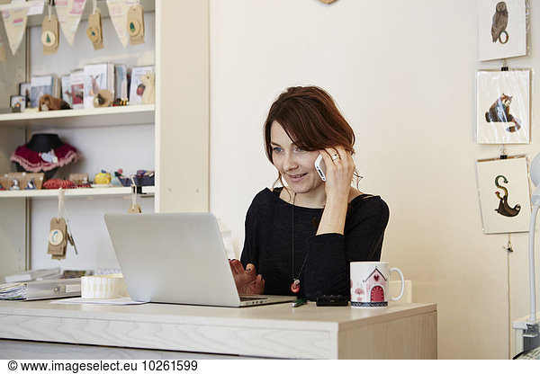 A woman sitting at a desk in a gift shop using a laptop and making a call.