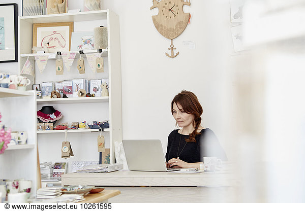 A woman sitting at a desk in a small gift shop  doing the paperwork  managing the business  using a laptop.