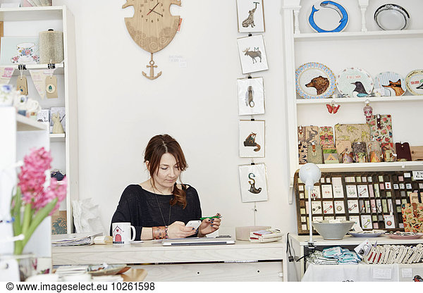 A woman sitting at a desk in a small gift shop  doing the paperwork  managing the business  using a laptop and a smart phone.