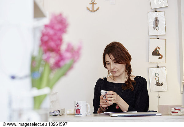 A woman sitting at a desk in a small gift shop  managing the business  making a call on a smart phone.