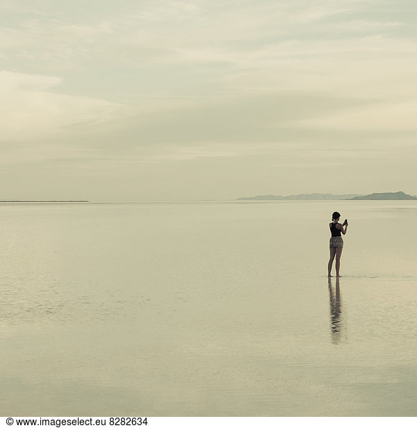 A Woman Standing On The Flooded Bonneville Salt Flats  Taking A Photograph With A Smart Phone.
