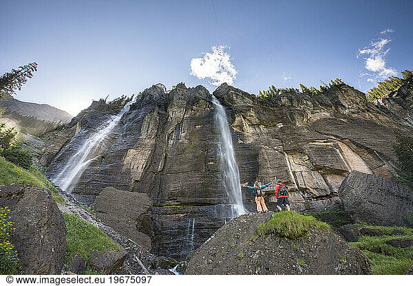 A woman stands with arms outstretched next to her hiking partner beneath a waterfall.