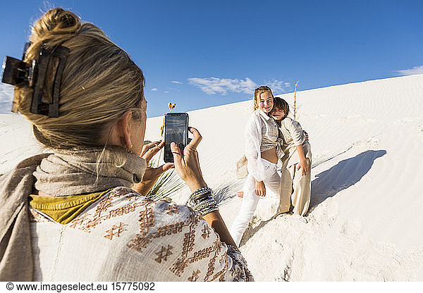 A woman taking picture of her children with a smart phone in white sand dunes landscape under blue sky.