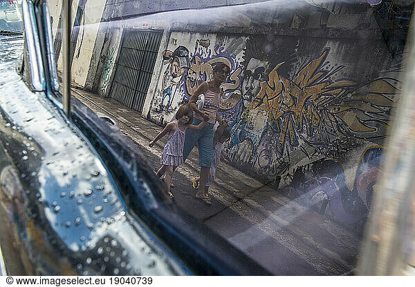 A woman walking with two young children  seen in the reflection of a window of a vintage car. Old Havana or La Habana Veija  La Habana  Cuba