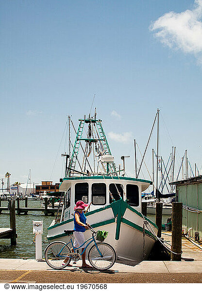 A woman wearing a pink floppy hat walks her bike in front of a fishing boat on a sunny day at a harbor.