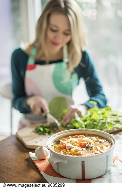 A woman wearing an apron  sitting at a table  chopping herbs  a bowl of vegetable stew in the foreground.