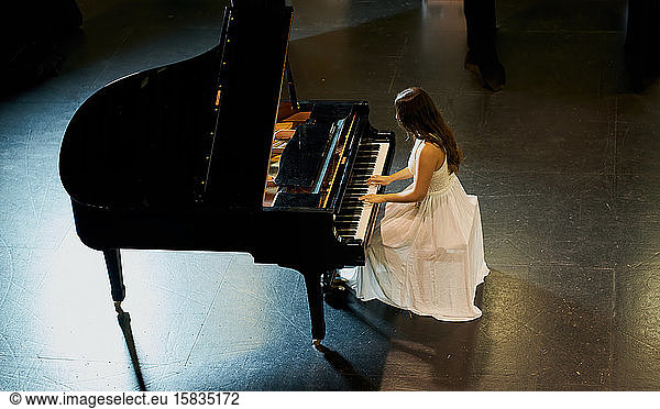 A woman with brown hair dressed in a white dress seen playing a black grand piano with the lid raised. View from above of the pianist