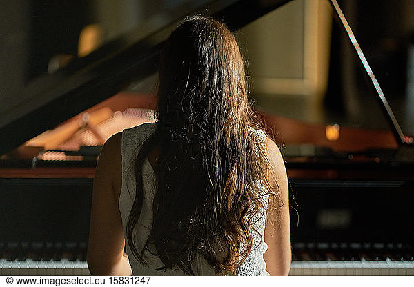 A woman with her back dressed in a white dress playing a black grand piano with the lid lifted