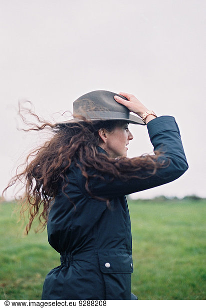 A woman with long hair being blown in the breeze  holding her hat.