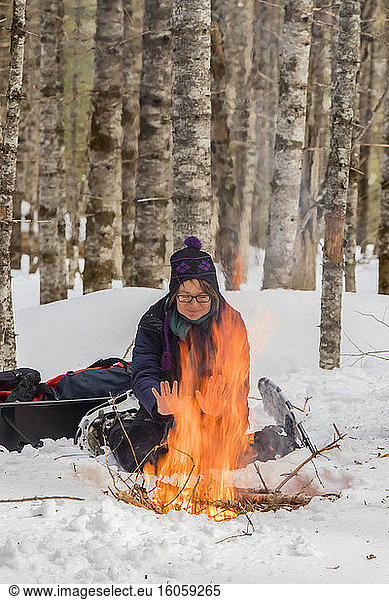 A woman with snowshoes sits on the snow and warms up by a campfire in a forest in wintertime  Gaspesie National Park; Quebec  Canada