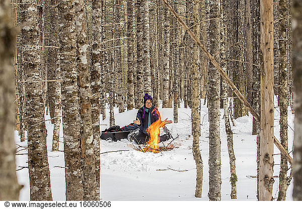 A woman with snowshoes sits on the snow and warms up by a campfire in a forest in wintertime  Gaspesie National Park; Quebec  Canada