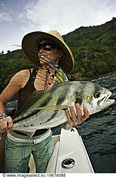 A women fishing in sunglasses and straw hat holds up a freshly caught rooster fish.