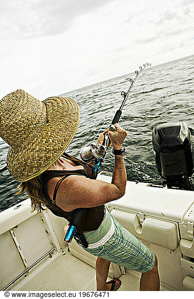 A women in a straw hat reels in a fish on the back of a boat with ocean in the background.