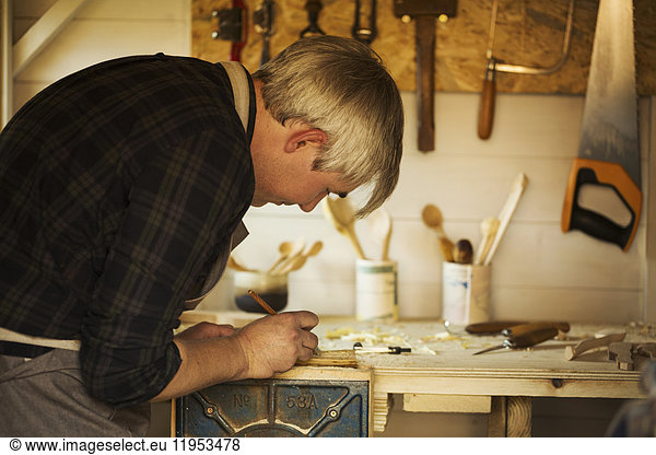 A wood carver craftsman  a wooden spoon carver at a workbench in his workshop  marking a piece of wood with a pencil.