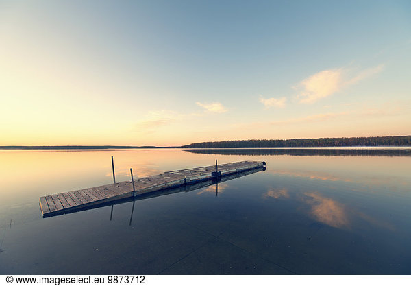 A wooden dock floating on flat calm waters of a lake at sunset.