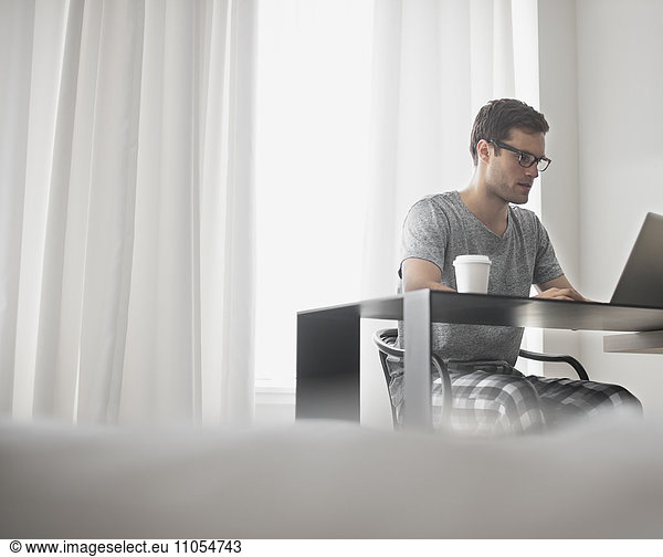 A working day. A man seated at a laptop computer  working in a hotel bedroom with a cup of coffee.