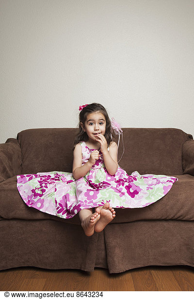 A 3 year old girl with long brown hair in a pink flowered cotton dress with the skirt spread out  sitting on a brown sofa.