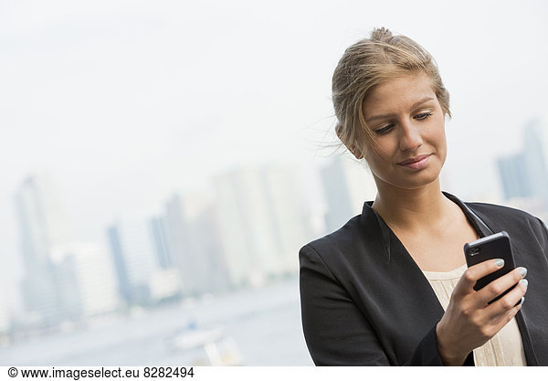 A Young Blonde Businesswoman On A New York City Street. Wearing A Black Jacket. Using A Smart Phone.