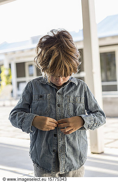 A young boy buttoning up his denim shirt
