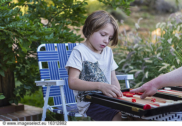 A young boy playing backgammon outdoors in a garden.