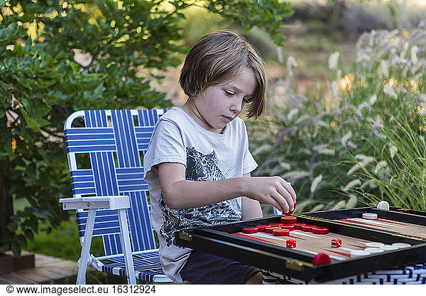 A young boy playing backgammon outdoors in a garden.
