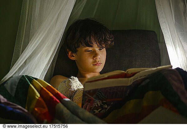 a young boy sits in chair with stuffed toy reading book at bedtime