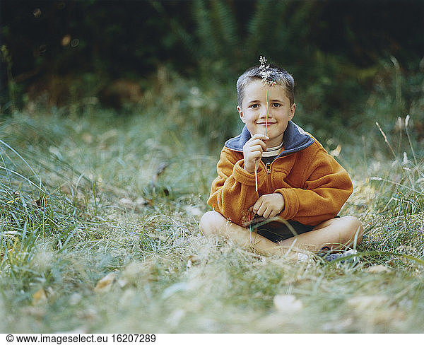 A young boy sits in field of tall grass  holding blade of grass