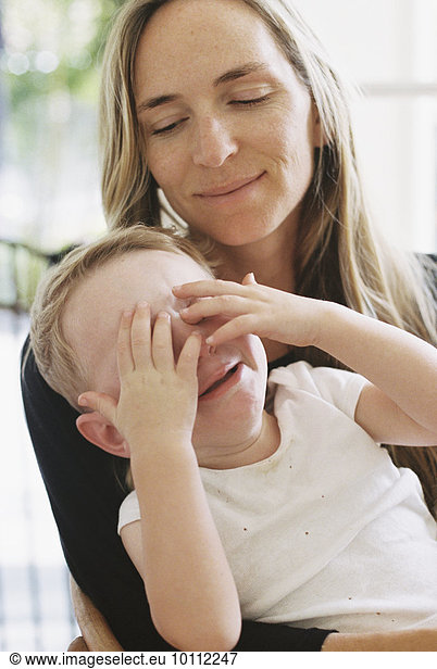 A young boy sitting on his mothers lap with his hands over his eyes.