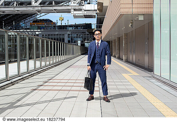 A young businessman in the city  on the move  a man in a suit with a laptop bag  standing legs apart on a walkway.