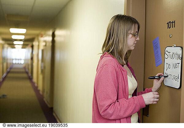 A young caucasian college student writes a note to her friends on the white board on her dorm door.