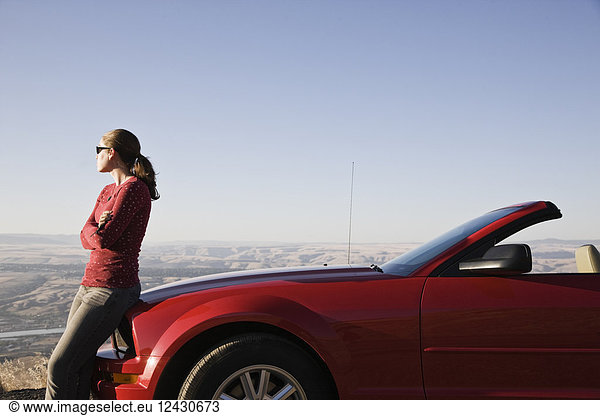 A young Caucasian woman leaning on her convertible sports car at a rest stop.